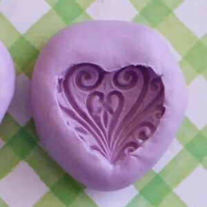 May include: A lavender silicone mold with a heart-shaped design and the text "Mold C". A white heart-shaped casting is also visible. The design features swirling patterns and a central floral element. The background is a green and white checkered pattern.