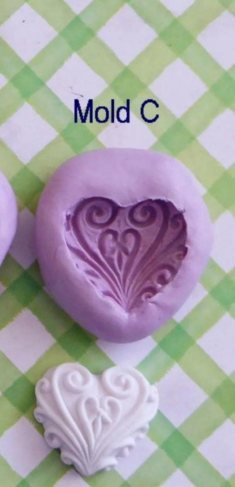 May include: A lavender silicone mold with a heart-shaped design and the text "Mold C". A white heart-shaped casting is also visible. The design features swirling patterns and a central floral element. The background is a green and white checkered pattern.