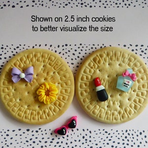 May include: Two 2.5-inch cookies with decorative elements. One cookie has a purple bow and yellow flower. The other has a lipstick and perfume bottle. Pink sunglasses with black hearts are also present. Text on the image reads: Shown on 2.5 inch cookies to better visualize the size.