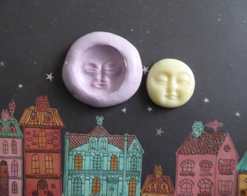 May include: A lavender silicone mold and a yellow moon-shaped resin piece with closed eyes. The mold is round with a recessed moon face. The resin moon has a smiling face. The background features a night sky with stars and colorful houses.