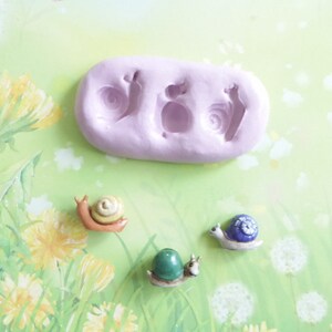 May include: A light purple silicone mold with three snail impressions, alongside three miniature snail figurines. The snails have colorful shells in shades of yellow, blue, and green. The background features a floral design.