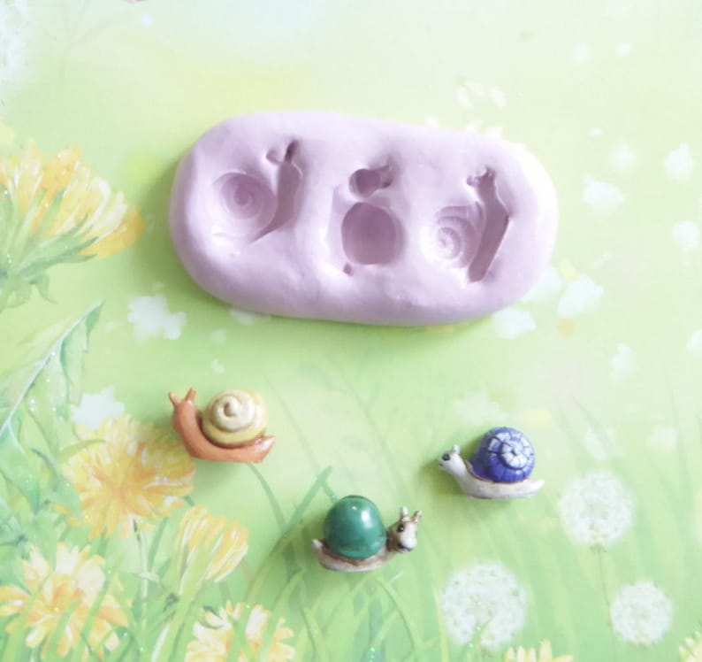 May include: A light purple silicone mold with three snail impressions, alongside three miniature snail figurines. The snails have colorful shells in shades of yellow, blue, and green. The background features a floral design.