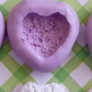 May include: A lavender heart-shaped mold with a floral pattern, labeled "Mold B". Below, a white heart-shaped object with a floral design is visible. The background features a green and white checkered pattern.