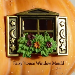 May include: A miniature bronze-colored window with open shutters, adorned with stars and crescent moons. The window box is filled with green leaves and purple and orange flowers. The text "Fairy House Window Mould" is below the window.