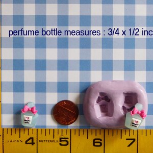 May include: A light blue perfume bottle miniature with a pink bow and heart detail, next to a penny and a mold. The text reads "perfume bottle measures: 3/4 x 1/2 inch". The background is a blue and white checkered pattern.