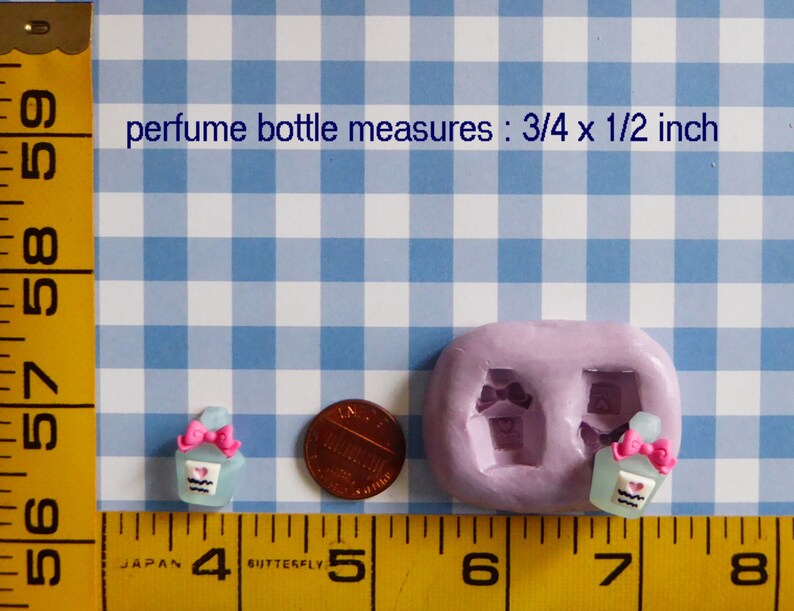 May include: A light blue perfume bottle miniature with a pink bow and heart detail, next to a penny and a mold. The text reads "perfume bottle measures: 3/4 x 1/2 inch". The background is a blue and white checkered pattern.