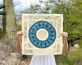 May include: A square textile piece featuring a central blue circular motif with intricate white patterns, surrounded by a cream-colored border with a geometric lattice design. The square fabric is held up, displaying a detailed blue and white print.