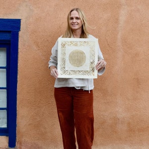 May include: A woman in a white shirt and brown pants is holding a square piece of art with a circular design in the center. The art is made of a woven material and has a light brown color.