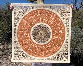 May include: A large, intricately cut paper artwork featuring a central circular design in shades of orange and beige. The artwork is held up against a backdrop of a blue sky and desert vegetation, including a tall cactus.