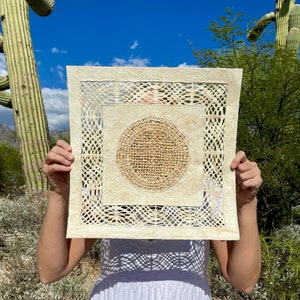 May include: A square piece of paper art with a circular design in the center. The paper is cream colored and has a woven texture. The circular design is made of a darker brown material and has a woven texture. The art is being held in front of a person's face.