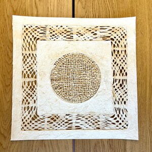 May include: A white square paper mat with a decorative, intricate, cut-out design. The mat features a circular design in the center with a woven texture.