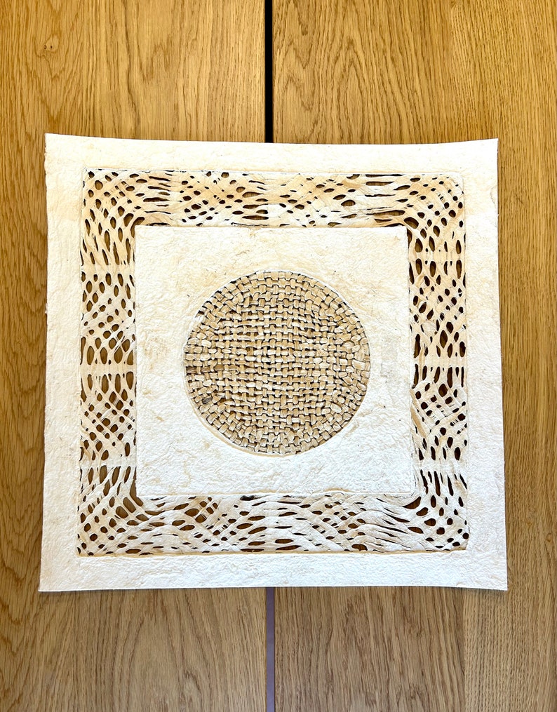 May include: A white square paper mat with a decorative, intricate, cut-out design. The mat features a circular design in the center with a woven texture.