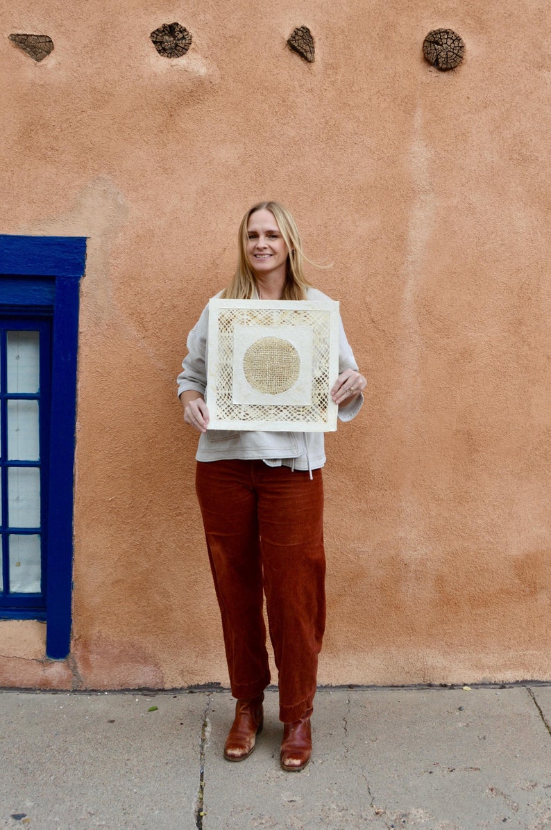 May include: A woman in a white shirt and brown pants is holding a square piece of art with a circular design in the center. The art is made of a woven material and has a light brown color.