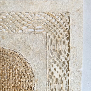 May include: A square wall hanging made of woven paper with a circular woven design in the center. The paper is a light beige color and the woven design is a darker beige color.
