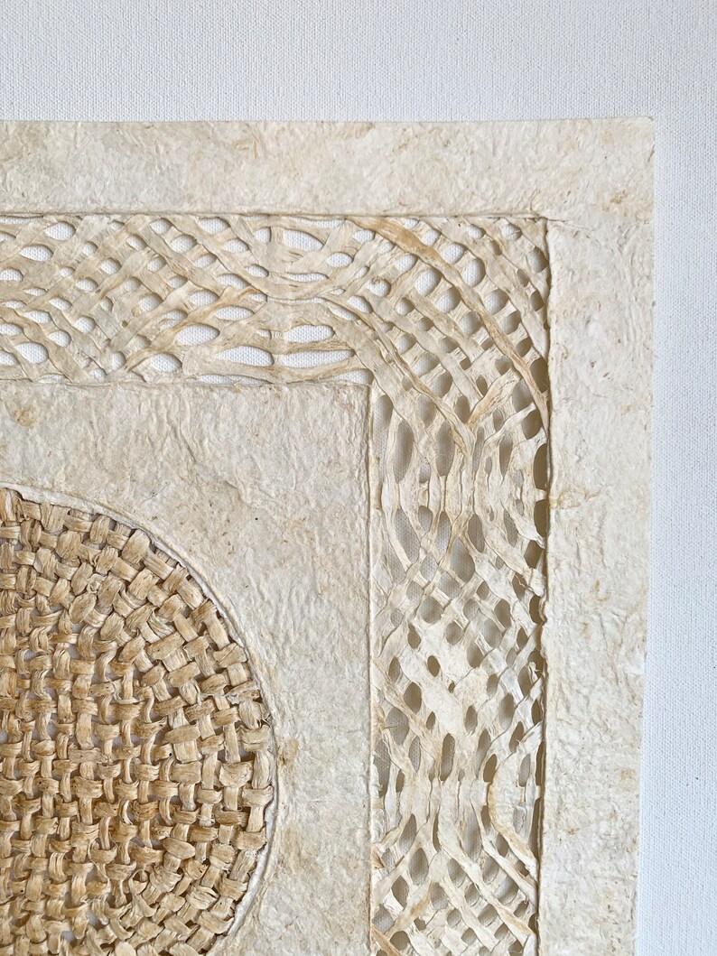 May include: A square wall hanging made of woven paper with a circular woven design in the center. The paper is a light beige color and the woven design is a darker beige color.