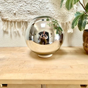 May include: A large, reflective silver orb on a white base, reflecting the surrounding room. The orb sits on a light-coloured wooden surface, with a woven wall hanging and potted plants in the background. The orb is a decorative item.
