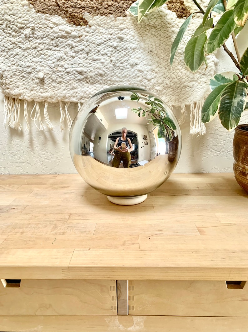 May include: A large, reflective silver orb on a white base, reflecting the surrounding room. The orb sits on a light-coloured wooden surface, with a woven wall hanging and potted plants in the background. The orb is a decorative item.