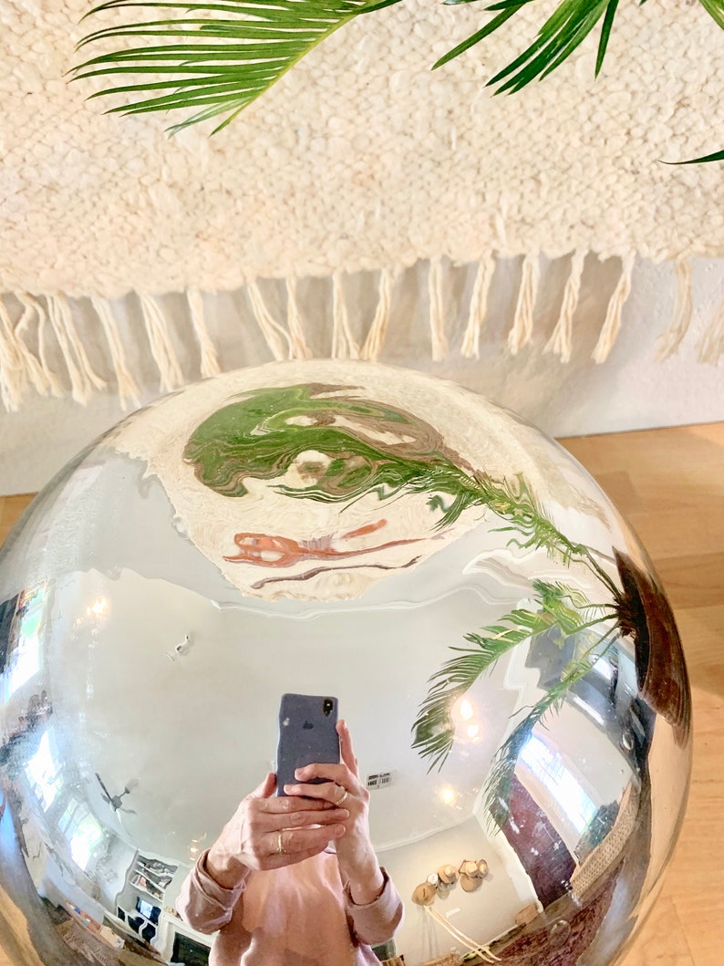 May include: A large, reflective silver sphere, reflecting a room with a rug, plants, and a person holding a phone. The sphere's surface is highly polished, creating a distorted mirror effect. Green palm fronds are visible.