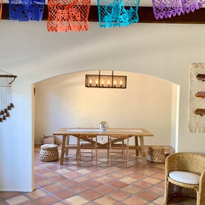 May include: A dining room featuring a wooden table, chairs, and woven baskets. The ceiling is adorned with colourful paper banners. The room has a warm atmosphere with terracotta tile flooring and a decorative light fixture.