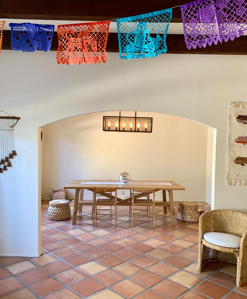 May include: A dining room featuring a wooden table, chairs, and woven baskets. The ceiling is adorned with colourful paper banners. The room has a warm atmosphere with terracotta tile flooring and a decorative light fixture.