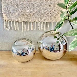 May include: Two silver, spherical decorative vases of different sizes sit on a wooden surface. The vases reflect the surrounding environment. A woven wall hanging with fringe hangs in the background.