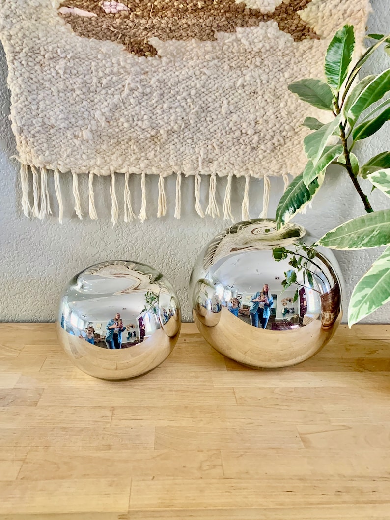 May include: Two silver, spherical decorative vases of different sizes sit on a wooden surface. The vases reflect the surrounding environment. A woven wall hanging with fringe hangs in the background.