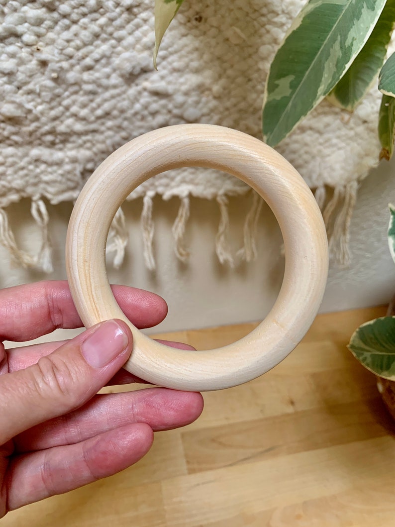 May include: A light-coloured wooden ring, held in a hand, with a smooth, unfinished surface. The ring is circular and appears to be made of a light-coloured wood. The background includes a textured, off-white fabric and green-leafed plants.