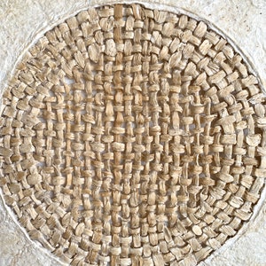 May include: A close-up of a woven circular design, made of natural fibers, with a light brown color. The woven design is set against a white background.