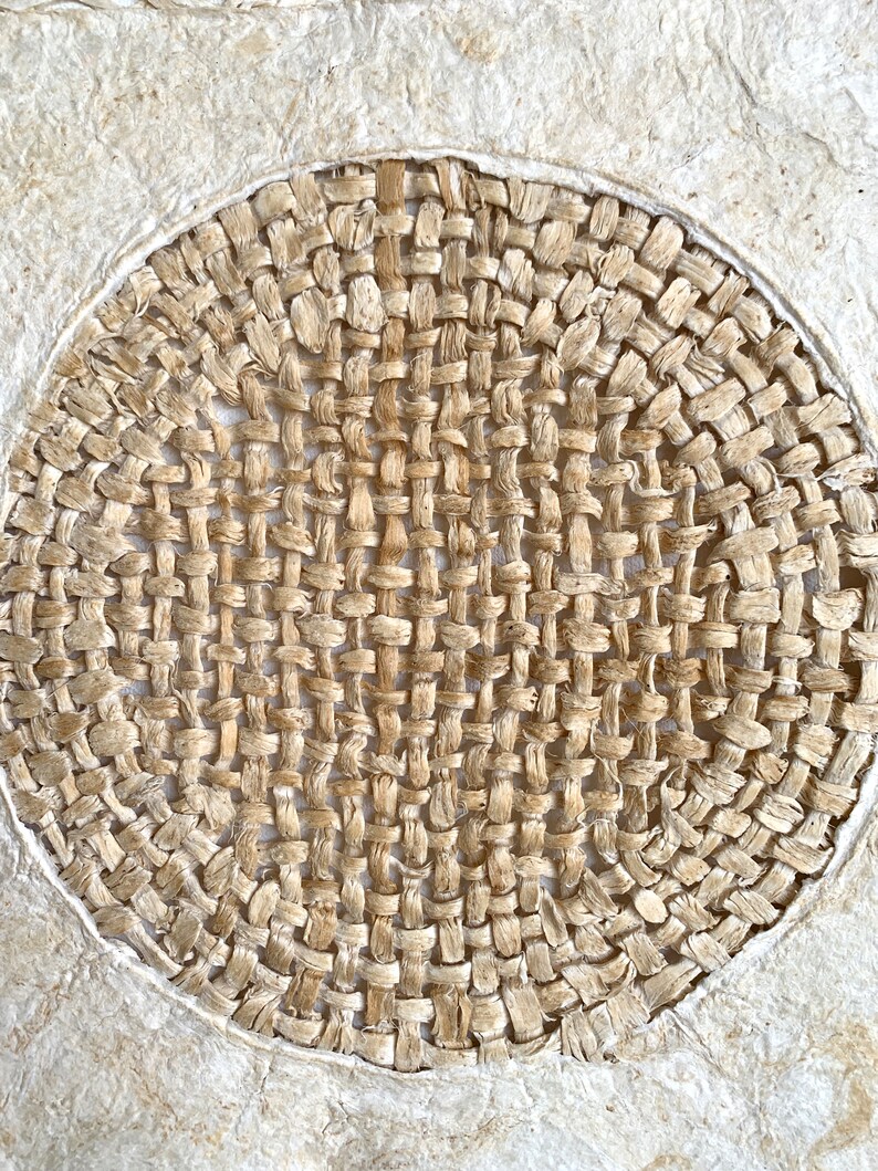 May include: A close-up of a woven circular design, made of natural fibers, with a light brown color. The woven design is set against a white background.