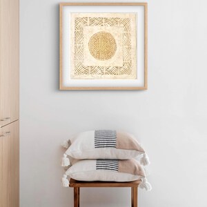May include: A framed artwork with a beige background and a woven circle design in the center. The artwork is hanging on a white wall above a wooden stool with two beige and black striped pillows.