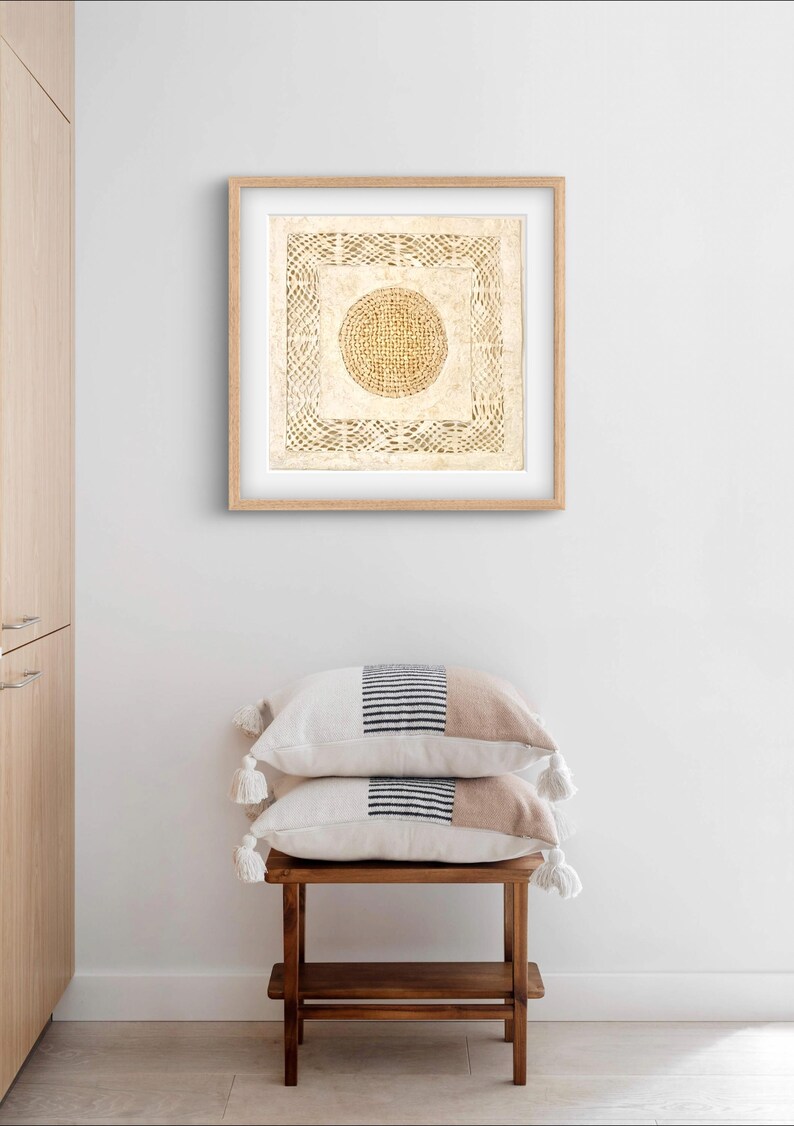 May include: A framed artwork with a beige background and a woven circle design in the center. The artwork is hanging on a white wall above a wooden stool with two beige and black striped pillows.