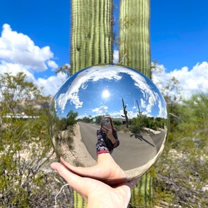 May include: A shiny metal sphere held in a hand, reflecting a sunny outdoor scene with a road, blue sky, and cacti. The sphere is the focal point, capturing the surrounding landscape.