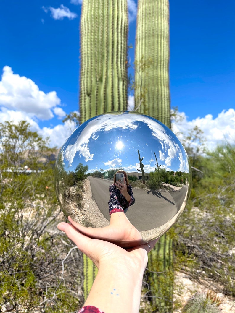 May include: A shiny metal sphere held in a hand, reflecting a sunny outdoor scene with a road, blue sky, and cacti. The sphere is the focal point, capturing the surrounding landscape.