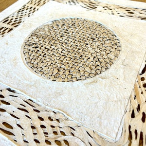 May include: A white paper with a woven design. The paper has a circular woven pattern in the center and a woven border around the edges.