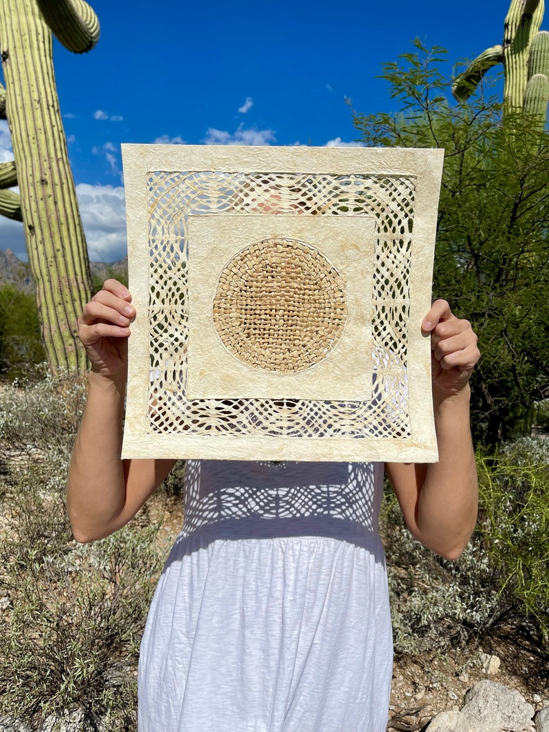 May include: A square piece of paper art with a circular design in the center. The paper is cream colored and has a woven texture. The circular design is made of a darker brown material and has a woven texture. The art is being held in front of a person's face.