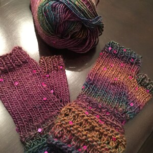 May include: Hand-knitted fingerless gloves in shades of purple, green, and brown, with pink sequin embellishments. A ball of matching yarn is next to them. The gloves have a ribbed cuff and are made with a textured stitch.