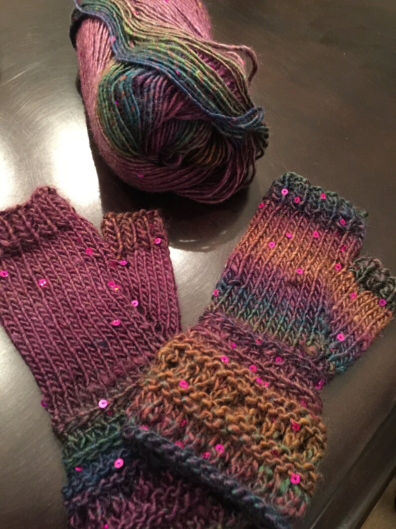 May include: Hand-knitted fingerless gloves in shades of purple, green, and brown, with pink sequin embellishments. A ball of matching yarn is next to them. The gloves have a ribbed cuff and are made with a textured stitch.