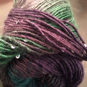 May include: A close-up of a ball of yarn with a gradient of colours. The yarn transitions from dark purple to teal, then to green, and finally to a grey-green. Small, silver sequins are scattered throughout the yarn, adding a touch of sparkle.