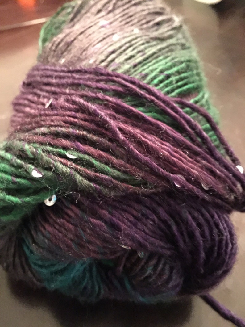 May include: A close-up of a ball of yarn with a gradient of colours. The yarn transitions from dark purple to teal, then to green, and finally to a grey-green. Small, silver sequins are scattered throughout the yarn, adding a touch of sparkle.