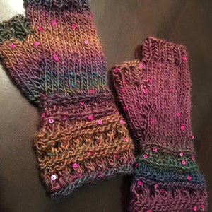 May include: Hand-knitted fingerless gloves in shades of purple, green, and brown, embellished with small, pink sequins. The gloves have a ribbed cuff and a textured pattern on the wrist. A cosy accessory for cooler weather.