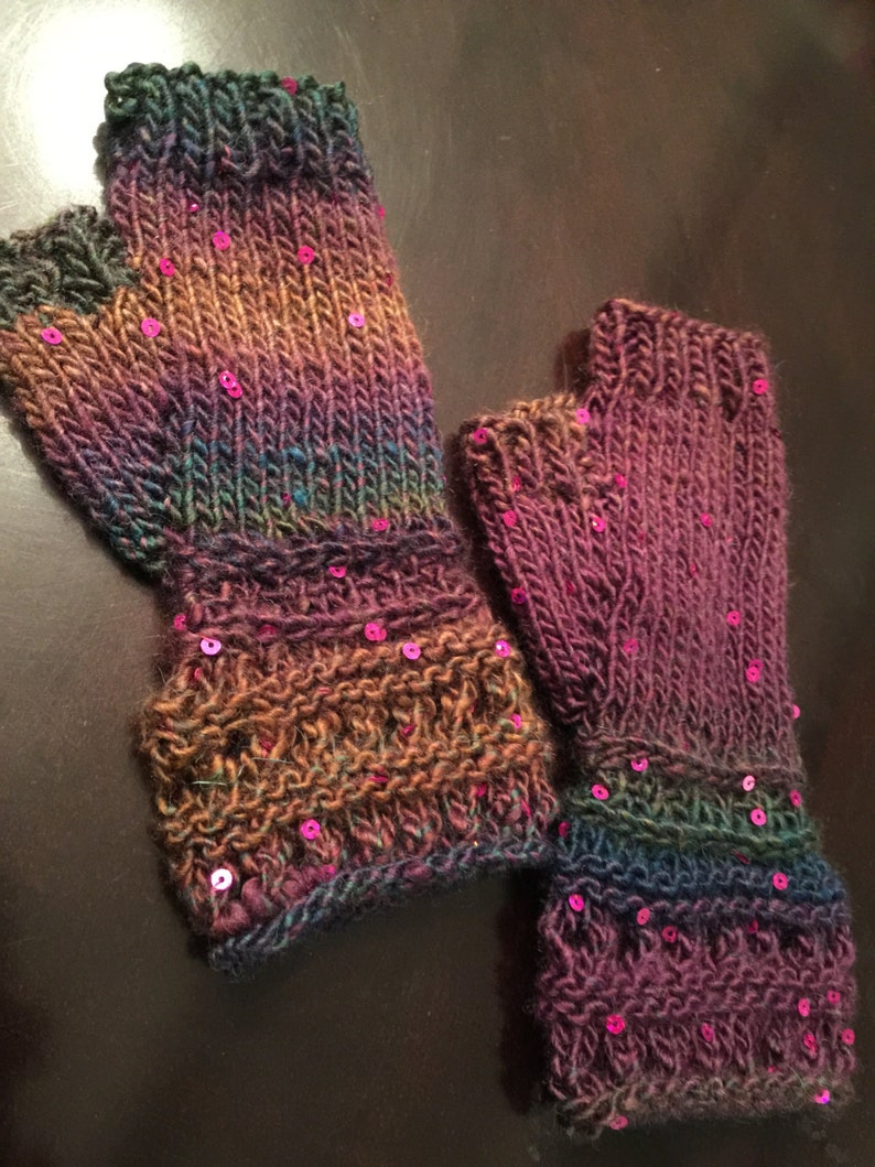 May include: Hand-knitted fingerless gloves in shades of purple, green, and brown, embellished with small, pink sequins. The gloves have a ribbed cuff and a textured pattern on the wrist. A cosy accessory for cooler weather.
