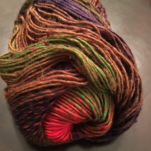 May include: A ball of hand-dyed yarn in a variety of colours. The yarn features shades of brown, green, purple, and red. The yarn is tightly wound and appears soft and textured, suitable for knitting or crochet projects.