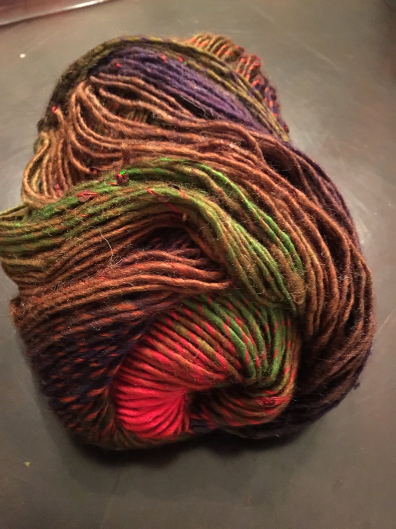 May include: A ball of hand-dyed yarn in a variety of colours. The yarn features shades of brown, green, purple, and red. The yarn is tightly wound and appears soft and textured, suitable for knitting or crochet projects.