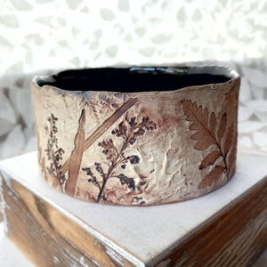 May include: A cylindrical ceramic bowl with a textured, light beige exterior featuring dark brown botanical leaf and stem imprints. The interior of the bowl is finished in a smooth, solid dark black glaze, creating a contrast with the earthy, organic patterns on the outer surface of the vessel.