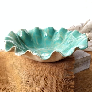 May include: A ceramic bowl with a ruffled, wavy edge and a glossy turquoise interior glaze. The exterior of the bowl features a textured, matte beige finish. The bowl has a wide, shallow basin shape with deep, organic folds along the rim.
