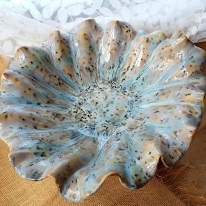 May include: A ceramic bowl with a ruffled, flower-like edge featuring a glossy glaze in shades of light blue, tan, and cream. The surface is decorated with small, dark brown speckles distributed throughout the interior and along the wavy rim.
