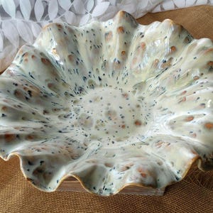 May include: A ceramic bowl with a ruffled, wavy edge and a glossy finish. The surface is cream-colored with a speckled pattern of small dark blue and light brown spots distributed across the interior and the undulating, flared rim.