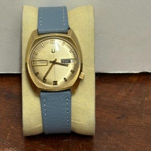 May include: A gold-toned Bulova Accutron watch with a light blue leather band. The watch face displays the day and date, with the Accutron logo at the bottom. The watch is displayed on a yellow cushion.