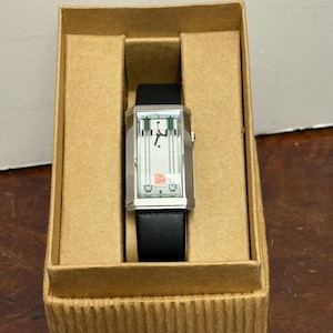 May include: A rectangular silver wristwatch with a black leather band, displayed in a cardboard box. The watch face has a white background with green and silver accents. The watch is labeled "Bulova".