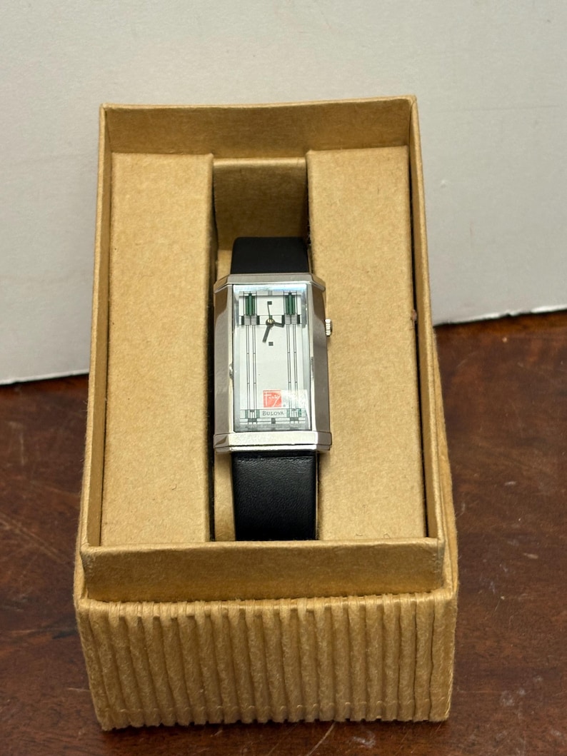 May include: A rectangular silver wristwatch with a black leather band, displayed in a cardboard box. The watch face has a white background with green and silver accents. The watch is labeled "Bulova".